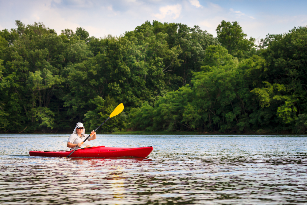 A woman kayoing on a lake with green trees in the background.