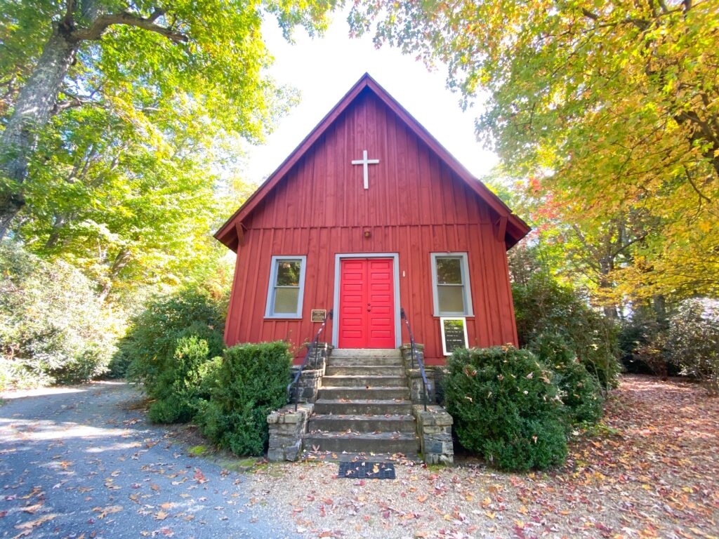 A small, red, wooden church surrounded by yellow trees and fallen leave.