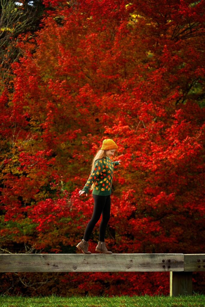 A woman in a leaf sweater balances on a fence in front of vivid red trees in North Carolina.