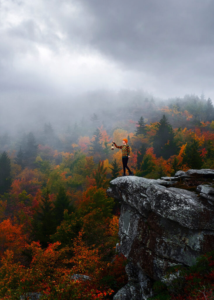 Woman holds an old fashioned lantern on a cliff overlooking a misty fall forest.
