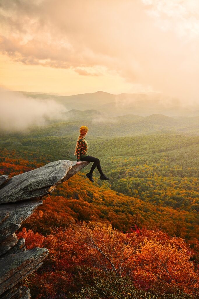 woman sitting on a rock ledge overlooking blue and yellow fall foliage below her wearing black pants and an orange top