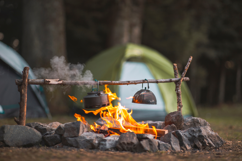 photo of two pans hanging on a stick roasting over a fire with a green tent in the background