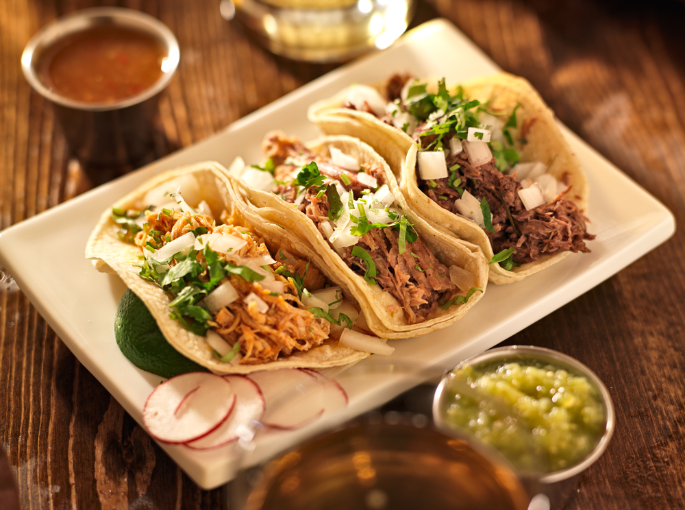 three tacos on a rectangle plate with salsas, radishes, and lime