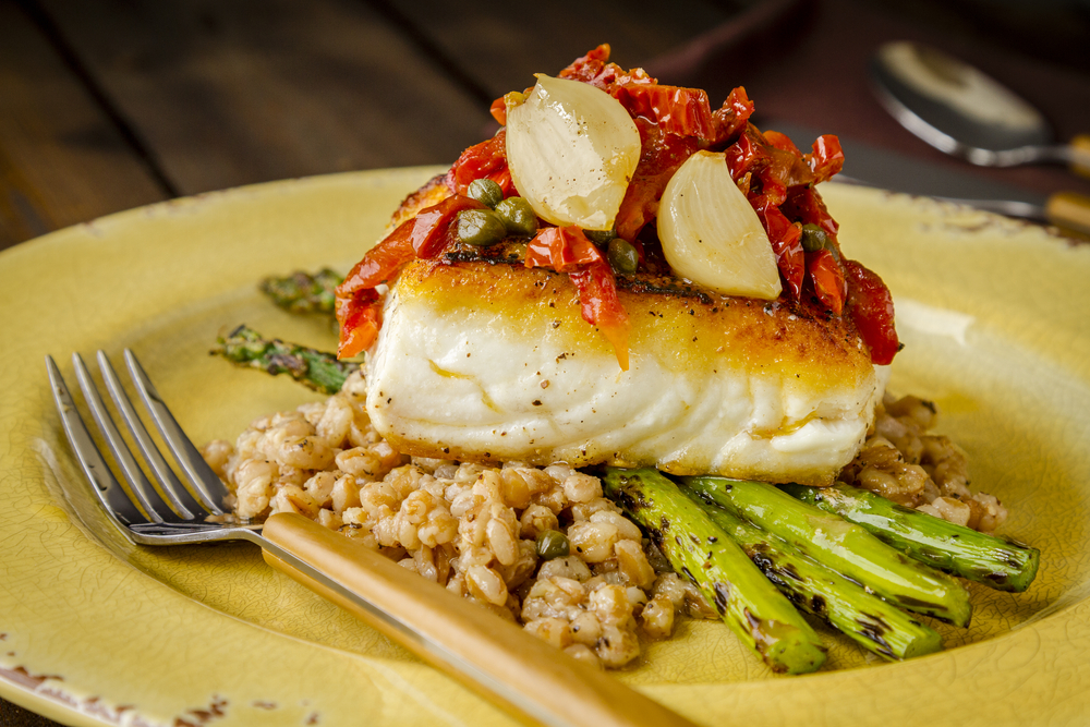 Dinner plate of fresh halibut fillet over bed farro with grilled asparagus, topped with salsa of red peppers, sundried tomatoes, garlic and capers on rustic yellow plate
