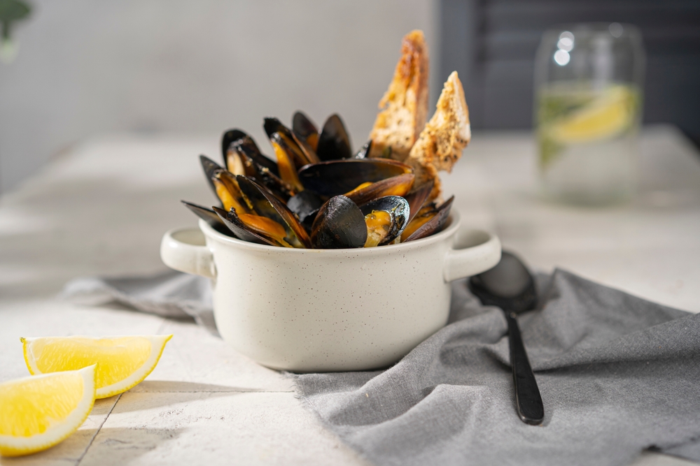 Cooked, boiled mussels with lemon juice, parsley and toasted bread in white bowl (plate), glass with fresh water on the table.