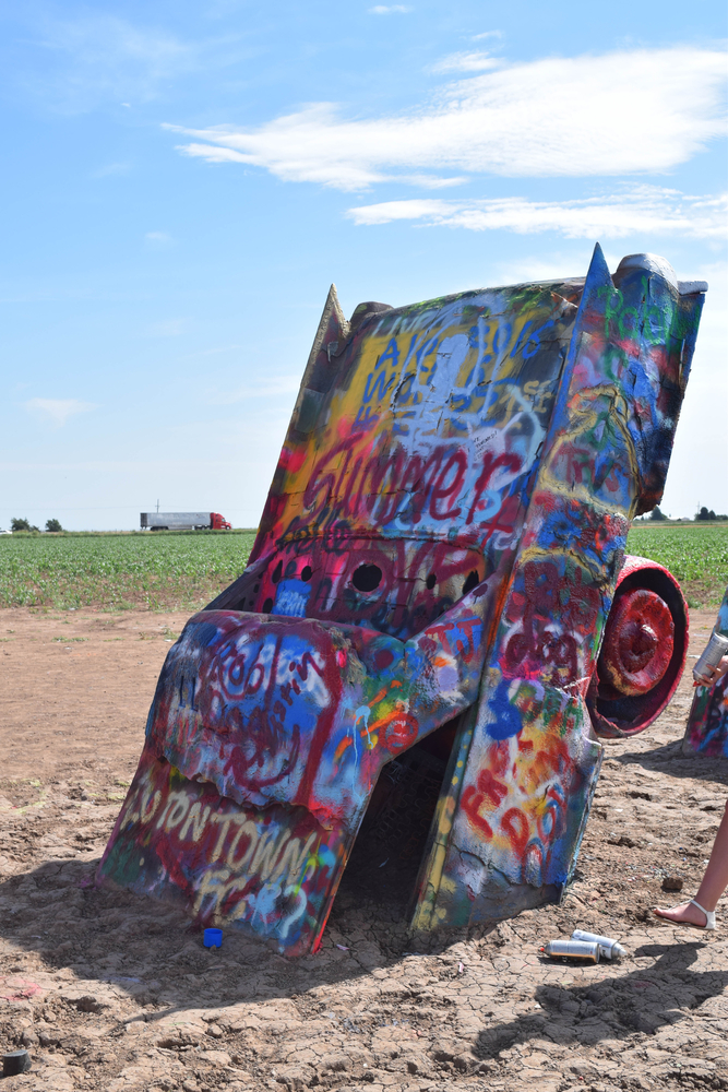 A Cadillac car sticking straight up out of the dirt with it's tail end pointing at the sky. It's covered in all kinds of graffiti in different colors. 