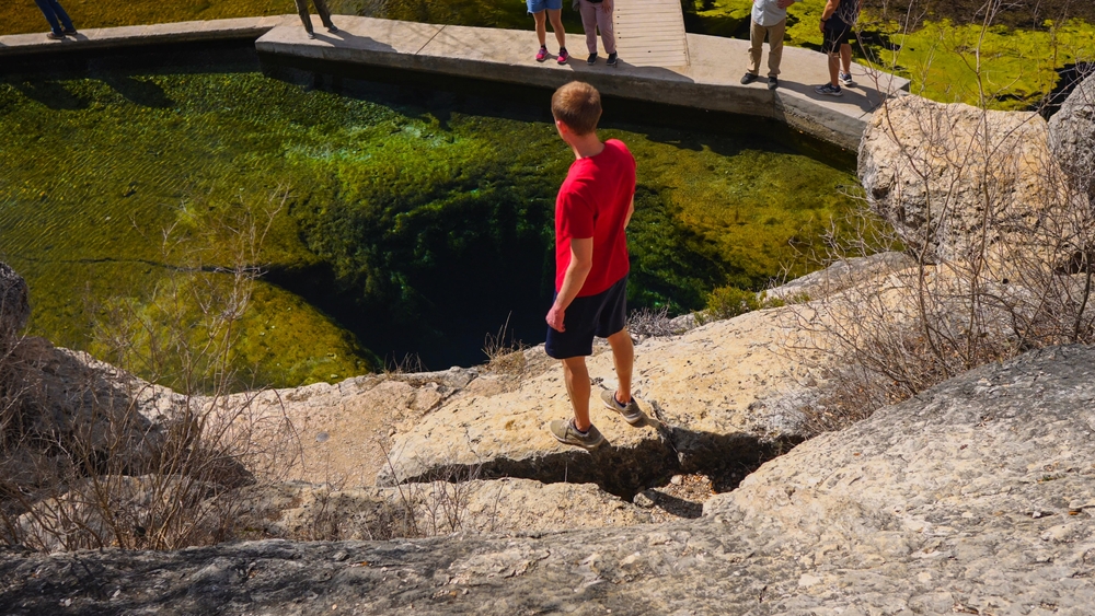 A person standing on a rock looking down into a deep well with crystal clear water one of the best hidden gems in Texas