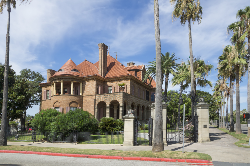 The exterior of a historic brick mansion in Galveston Texas, surrounded by palm trees, on a sunny day