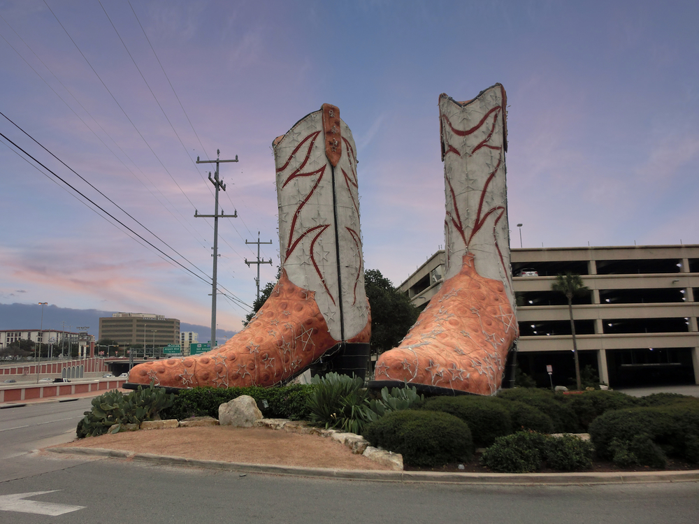 A sculpture that is a large pair of cowboy boots next to a parking garage as the sun is setting in the distance. 