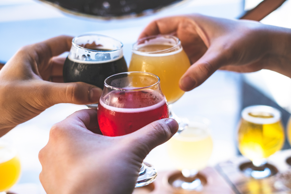 three hands holding three glasses of beers over a flight of beer on a table