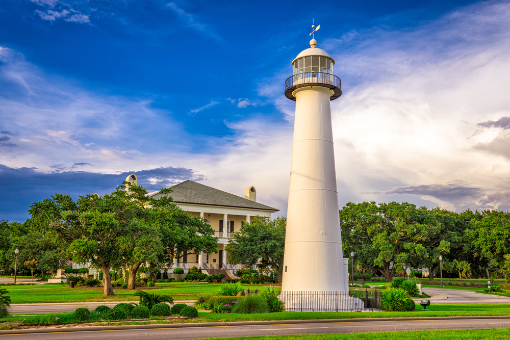The white Biloxi lighthouse stands against a blue sky surrounded by greenery, with the Biloxi Visitors Center in the background.
