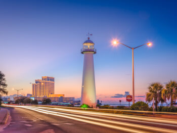 The Biloxi Lighthouse at sunset, with streetlights and headlights glowing, with the Beau Rivage Resort in the background.
