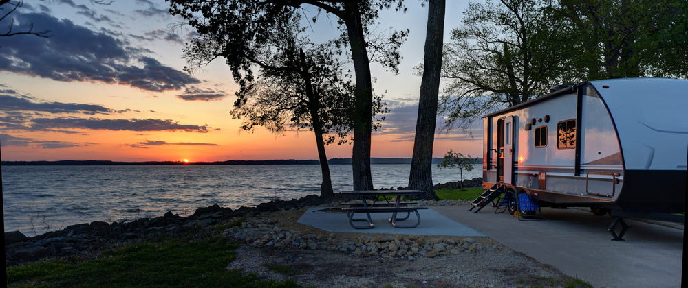 photo of an RV set up along the lake, overlooking a sunset at the far end of the lake with a picnic bench 