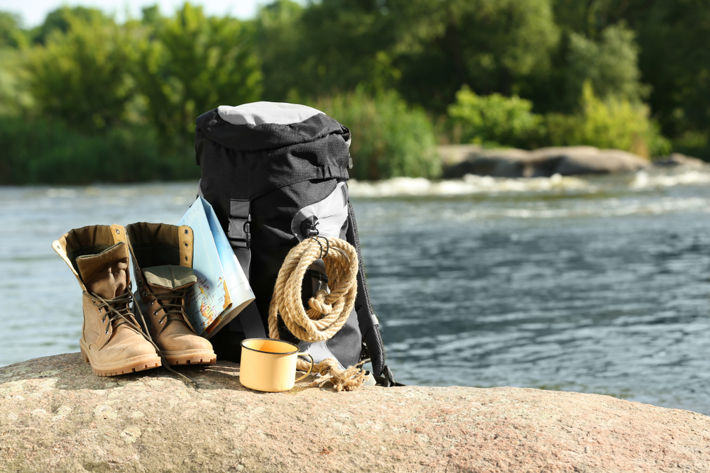 photo of a black backpack with a rope, hiking boots, a map and coffee mug sitting on a rock, with a river in the background 