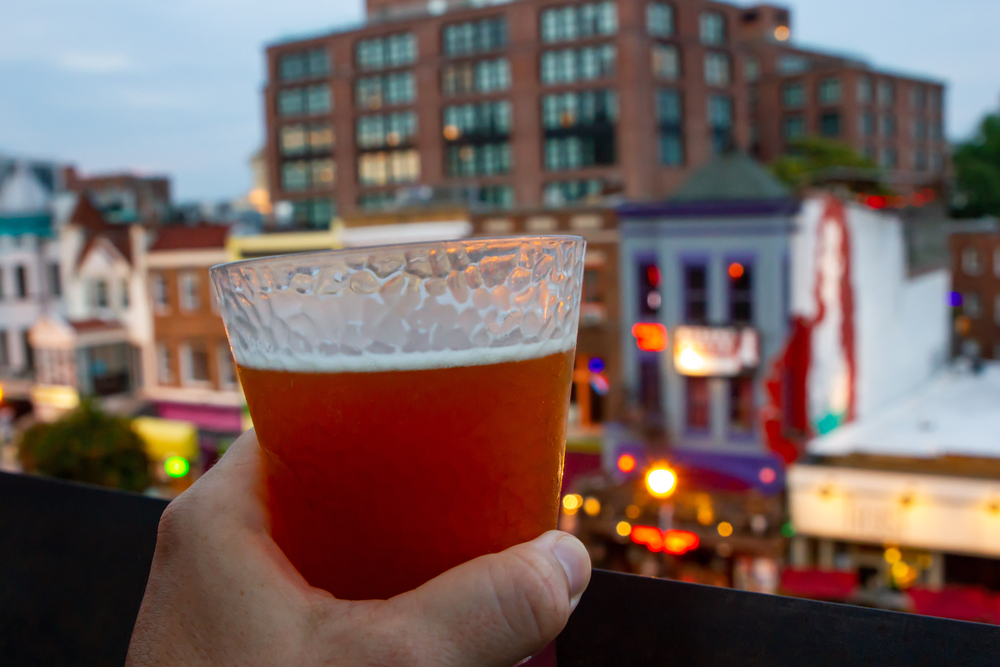 A hand holding a glass of beer with bars and restaurants lit up in the background as the sunsets