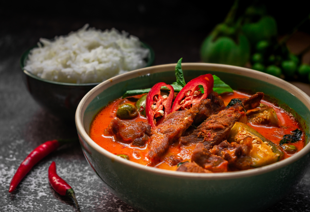 a big bowl of red Thai spicy soup with peppers and rice on the side. 