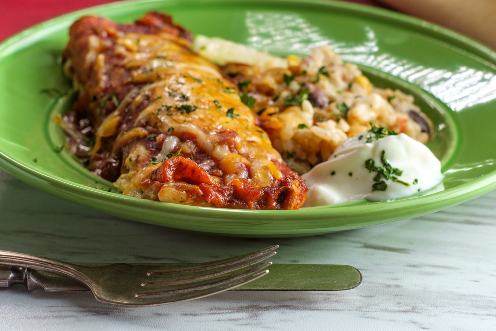 a plate next to a fork and knife, a enchilada, sauce, and rice and beans in the plate 