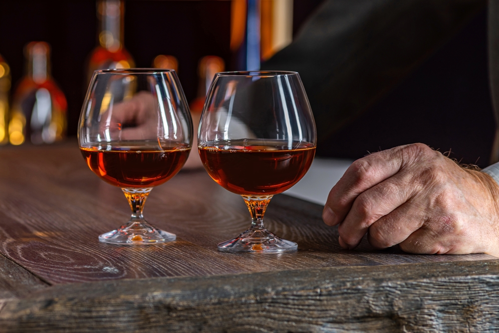 two glasses of rum sit on a bar, a mans hand is resting in the table beside the glasses. 