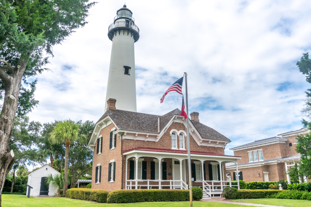 a brick building beneath a tall lighthouse, a flag pole is in front of the brick building with two flags on it 