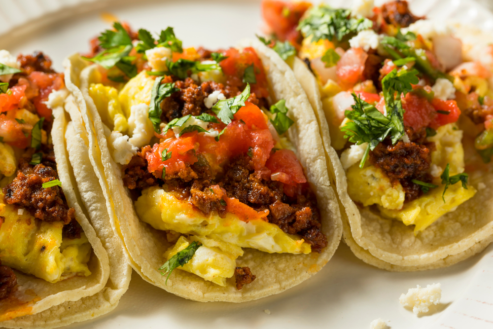 photo of three breakfast tacos on a white plate, filled with eggs, sausage, tomatoes and cilantro 