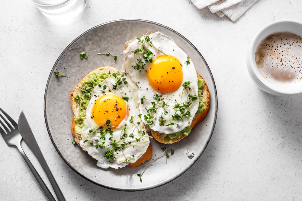 photo of two sunny side up eggs on toast with avocados and sprouts on a plate next to a cup of coffee 