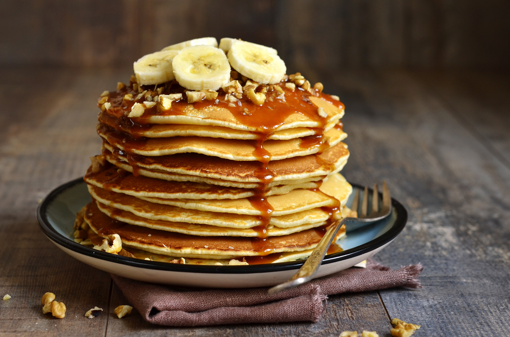 photo of a stack of pancakes with cut bananas on top with sprinkled walnuts and syrup dripping down the sides 