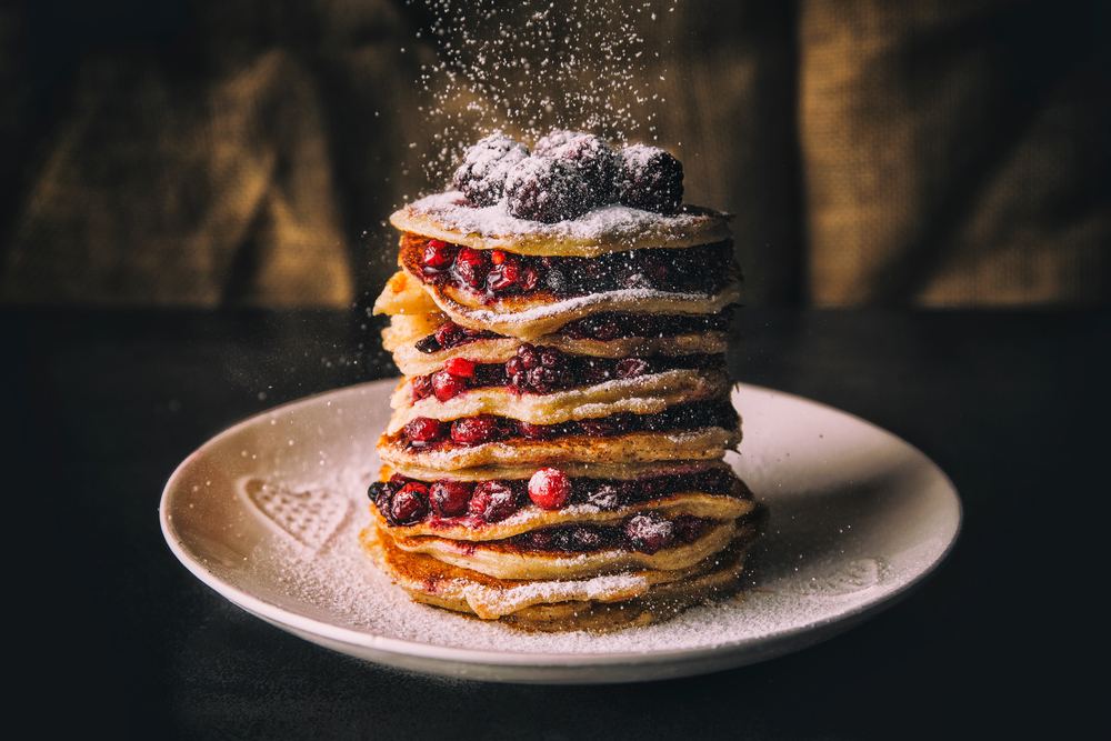 photo of a tall stack of pancakes with fruit layered between each pancake and powdered sugar sprinkling down 