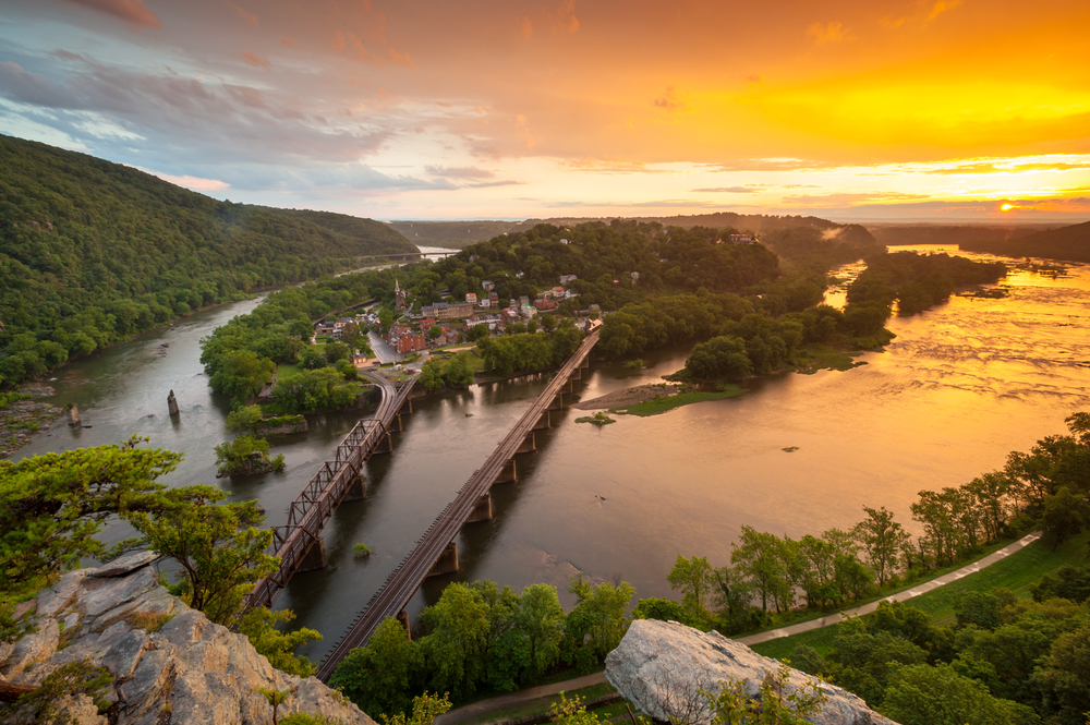 one of the best things to do in west virginia, harpers ferry, an arial image of an island between two rivers with two bridge leaving the mainland for the island at sunset