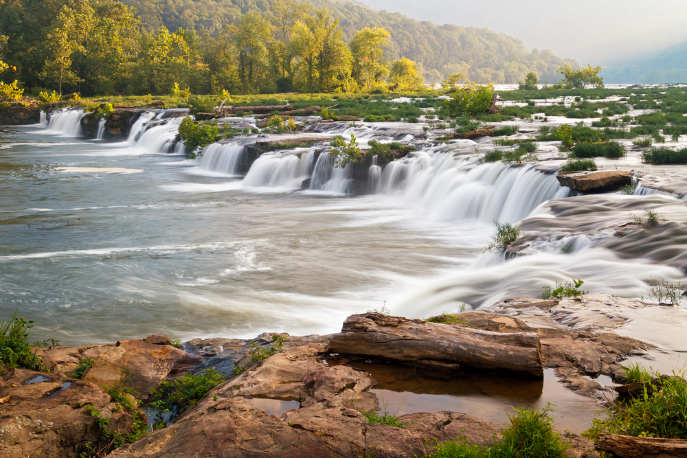 a long curved waterfall falling into a river, tree and mountains in the background, one of the best things to do in west virginia 