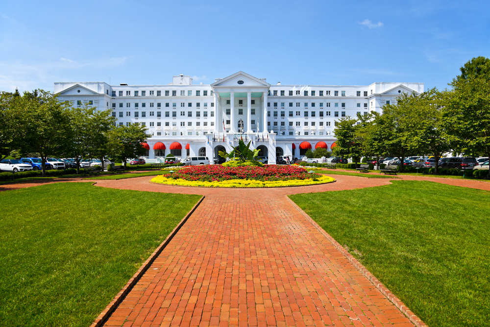 a tall and wide building with tree, grass, and brick walkway in front of it. A round flower garden sits in middle of brick walkway