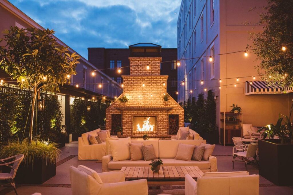 Outdoor lounge with white couches and string lights overhead with a roaring fireplace at dusk.