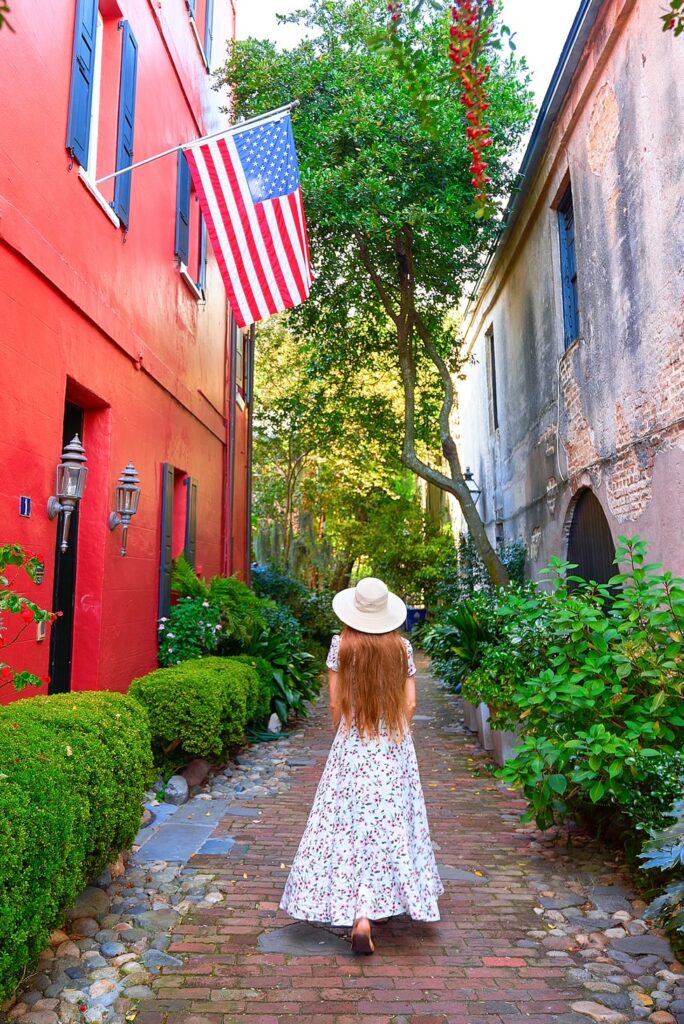 Woman in a floral dress and sun hat stands in Philadelphia Alley among shrubs under an American flag.