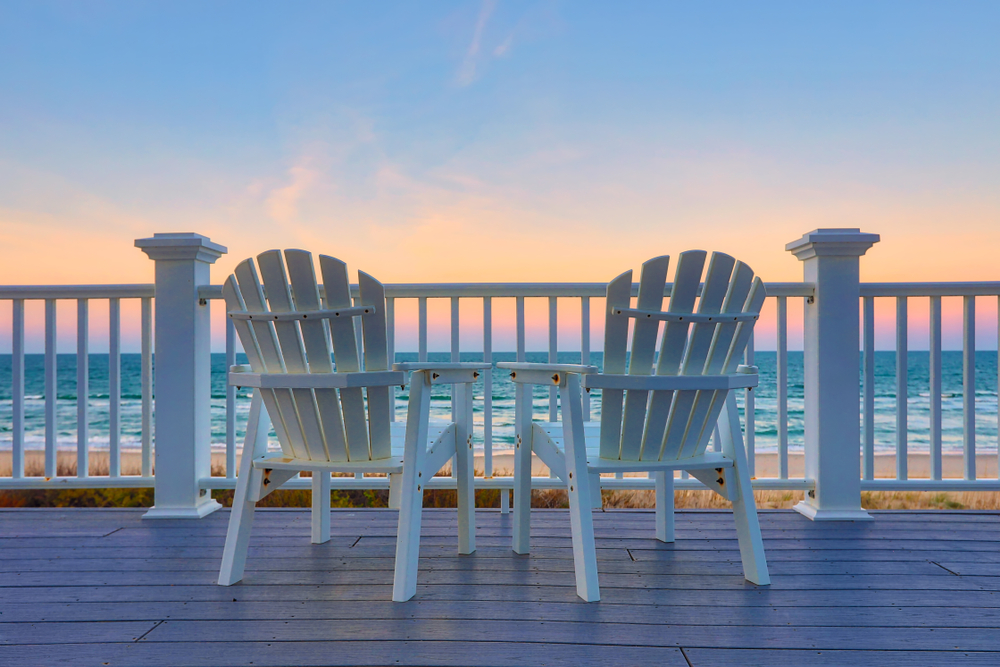 Two white Adirondack chairs on a deck overlooking the sunset over the ocean in the Outer Banks.