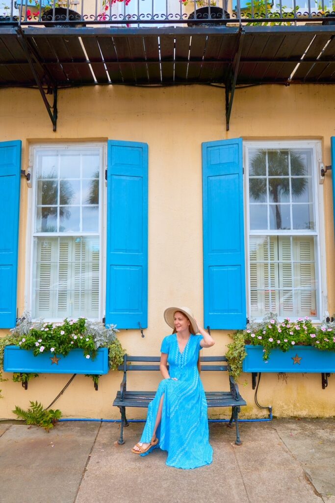Woman in blue dress and hat sitting on a bench in front of a yellow and blue house in Charleston, SC