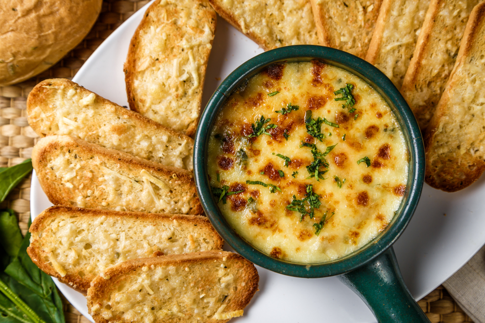 a small bowl with a handle, the bowl has crab dip in it and it surrounded by cheese bread for dipping