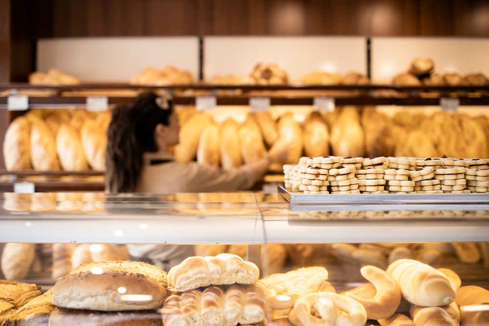 a woman gets bread from a shelf in a bakery, a tray of cookies is on the counter in the foreground