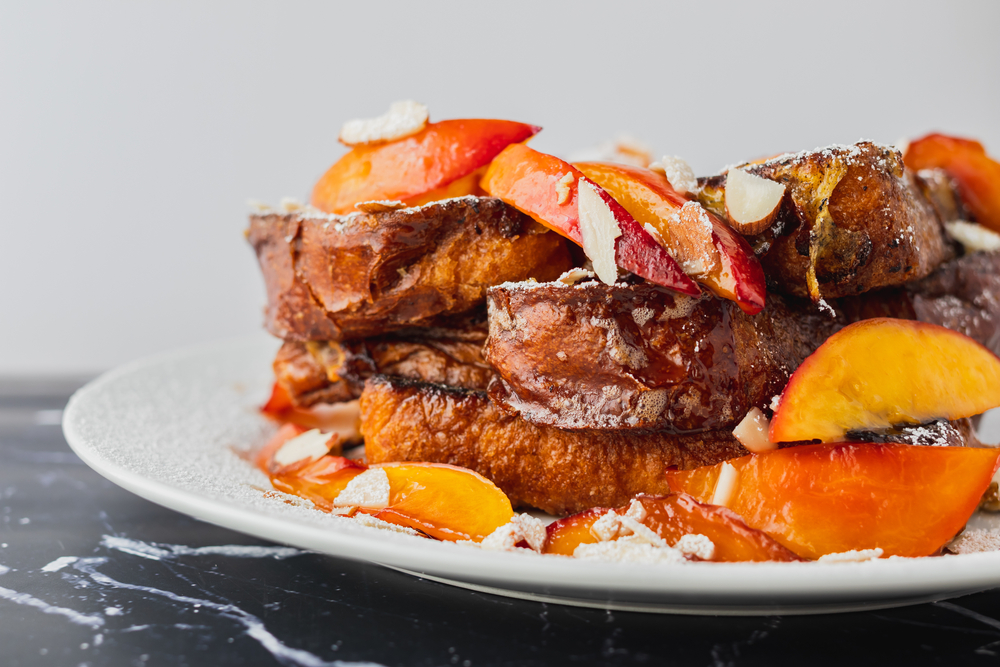 Peach cobbler French toast is fried and doused with syrup, almonds, powdered sugar, and peaches,