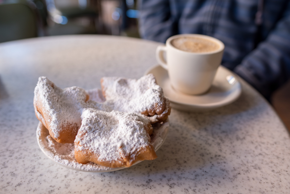 Three beignets covered in powered sugar sit on a plate next to a cup of coffee