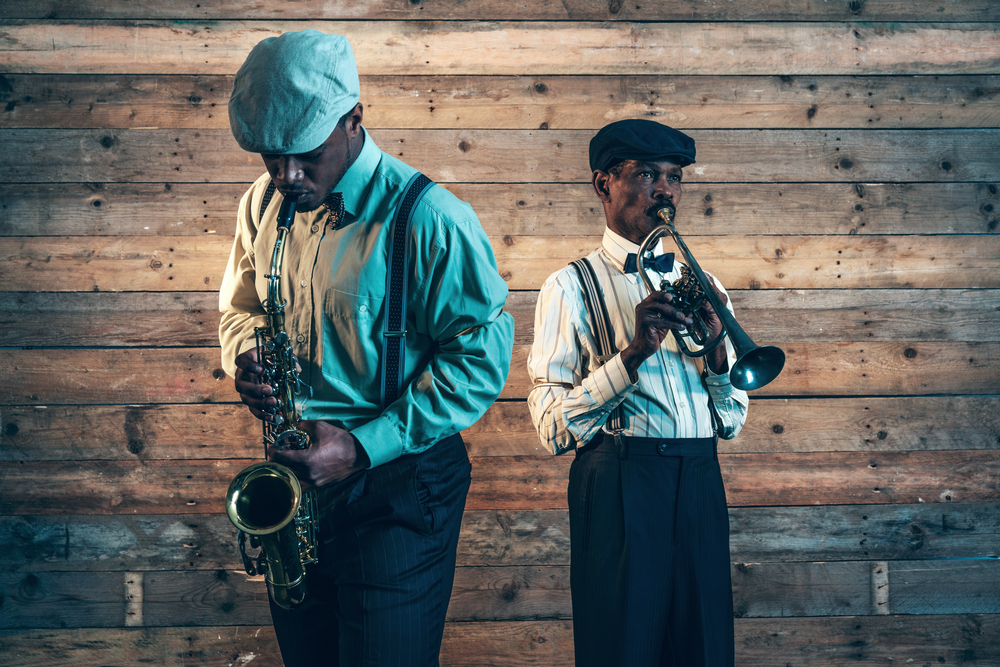 Two Black men playing the trumpet and the saxophone on a stage with a wooden wall behind them
