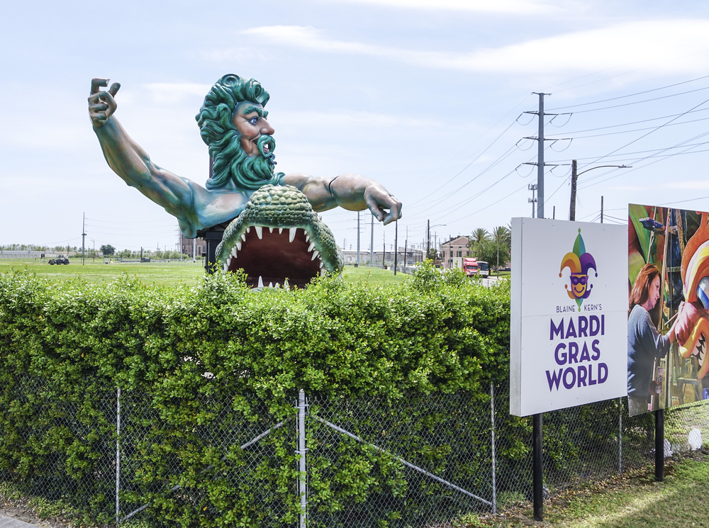 A large sculpture of a sea god and an alligator in a shrub by a sign for Mardi Gras World on a sunny day