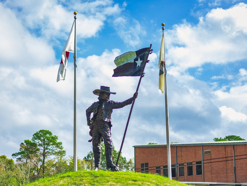 Photo of "The Texian" statue between two Texas flags on a sunny day at the Historic Flag Park in Conroe, TX