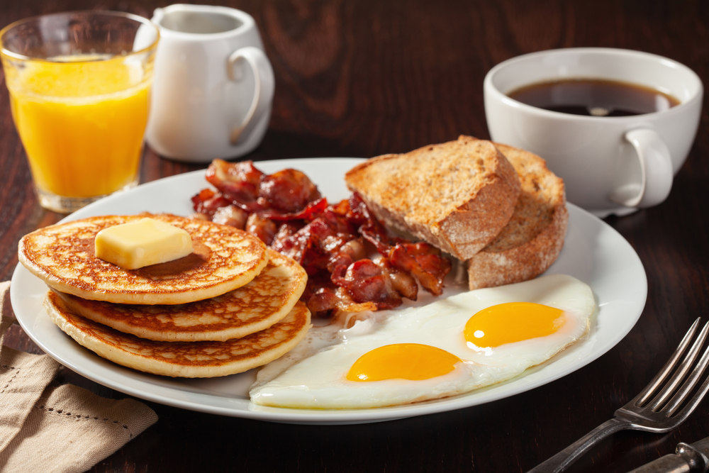 pancakes, bacon, toast, and eggs on a plate with a cup of coffee and a cup of orange juice on the table