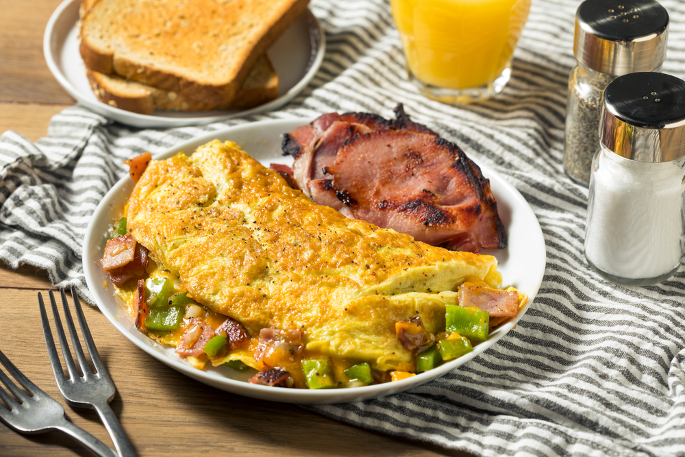 an omelete and ham on a plate next to a plate of toast, a glass of orange juice, and salt and pepper shakers