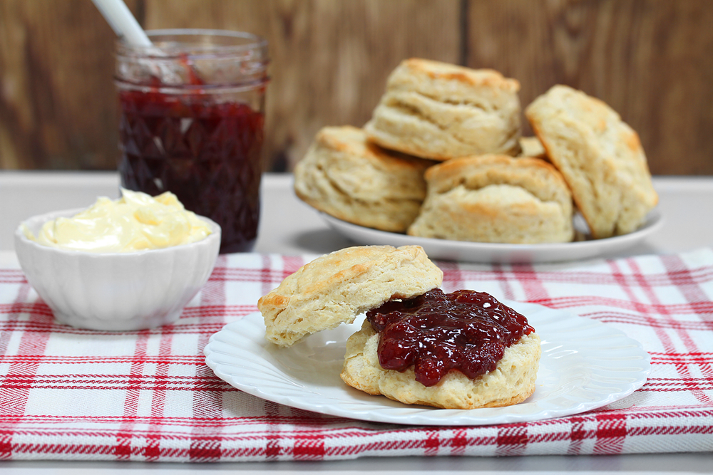 a large stack of biscuits on a plate, a jar of jam, and a bowl of butter sit next to a small plate that has a biscuit with jam on it