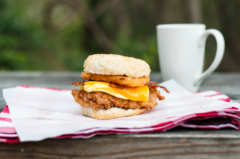 A chicken biscuit with an egg, fried chicken, and hashbrown sits on a biscuit with a cup of coffee in the background.