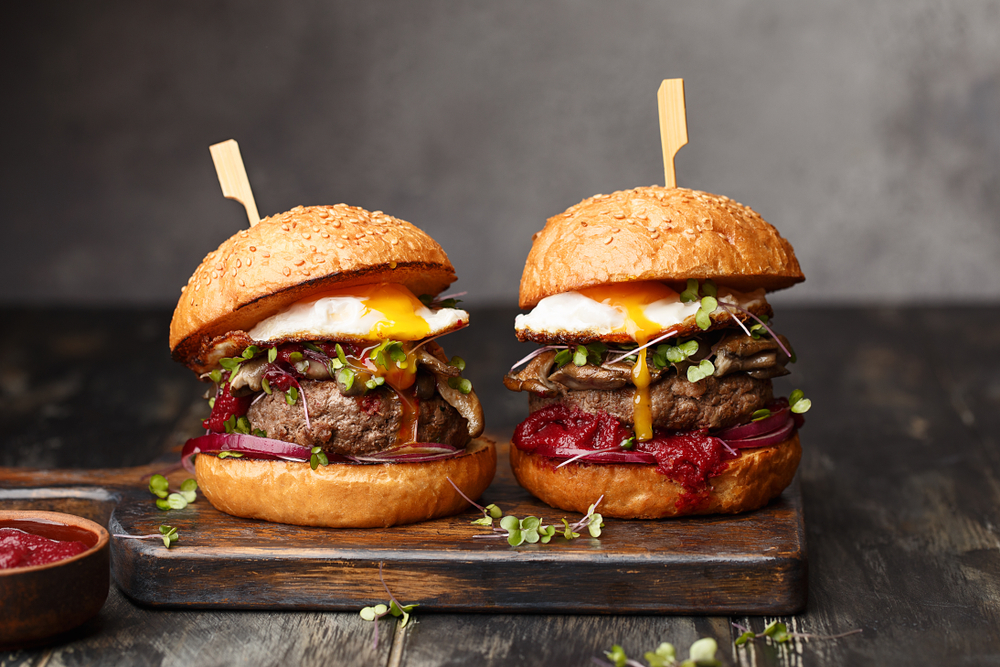 Two breakfast burger sliders sit on a slab of wood with running eggs dripping down the bacon and jam on the burger patty. 