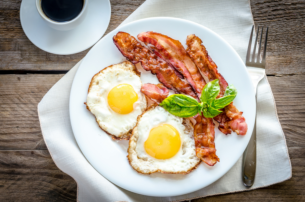 A plate features two sunny side up eggs, three strips of bacon, and a cup of black coffee on the side. 