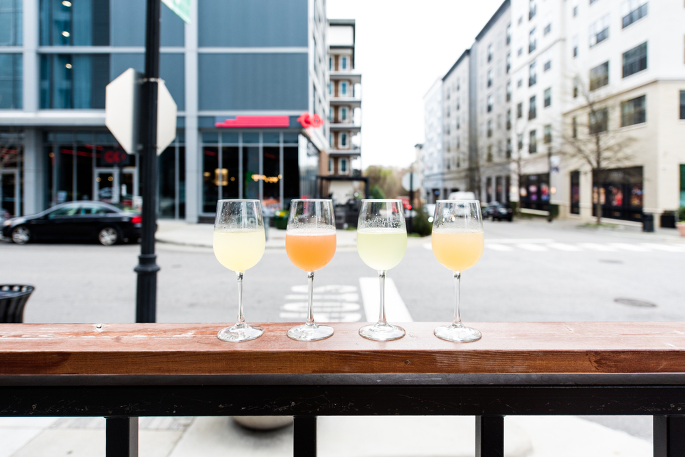 Four glasses sit on the edge of a bar, looking out over the city and down the road: these glasses contain four flavors of mimosas, making this mimosa flight a great option for taste testing.