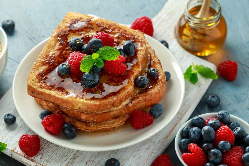 A stack of three slices of French toast are topped with syrup and blueberries and raspberries. 