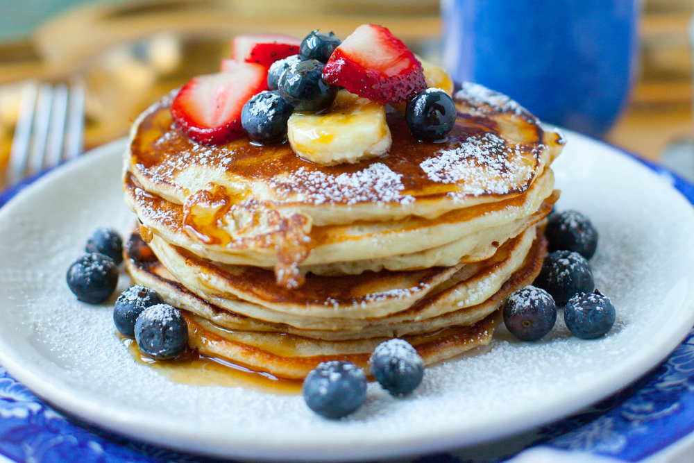 A stack of pancakes with fresh fruit and powdered sugars towers over a nice thin plate.